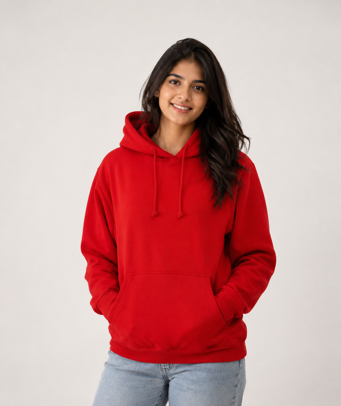 A woman wearing a bright red pullover hoodie with drawstrings and a front kangaroo pocket, paired with light blue jeans, standing against a plain light gray studio background.