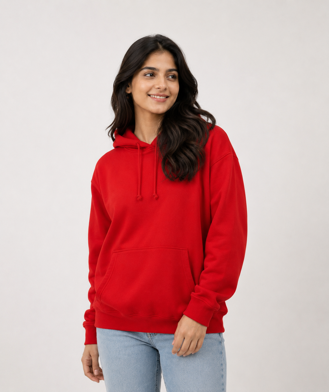 A woman wearing a bright red pullover hoodie with drawstrings and a front kangaroo pocket, paired with light blue jeans, standing against a plain light gray studio background.