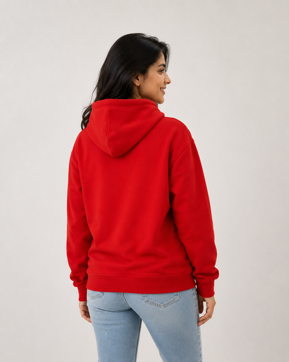 A woman shown from the back wearing a bright red pullover hoodie and light blue jeans, standing against a plain light gray studio background.