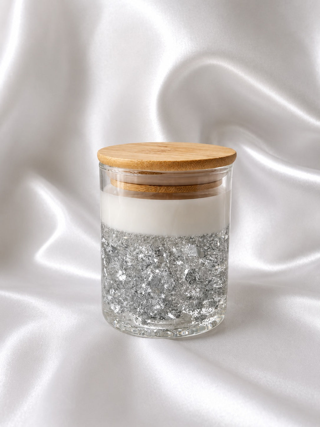 A clear glass jar with a light wood lid, filled with white wax and topped with silver glitter, set against a draped white satin background.