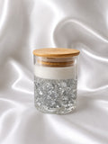 A clear glass jar with a light wood lid, filled with white wax and topped with silver glitter, set against a draped white satin background.