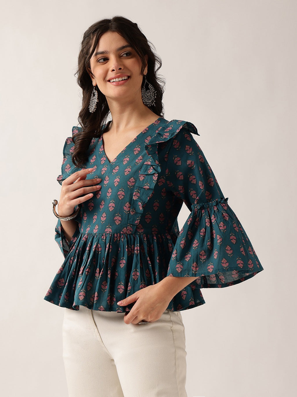 A woman modeling a dark teal green floral print blouse with ruffled shoulders and flared bell sleeves, paired with off-white pants, against a plain light beige background.