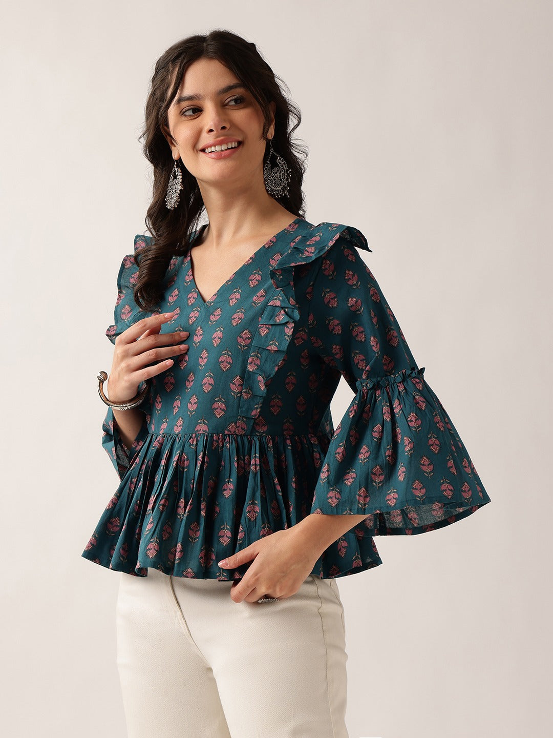 A woman modeling a dark teal green floral print blouse with ruffled shoulders and flared bell sleeves, paired with off-white pants, against a plain light beige background.