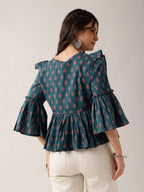 A woman wearing a teal green floral print blouse with ruffled sleeves and a peplum hem, paired with off-white trousers, shown from the back against a plain light gray studio background.