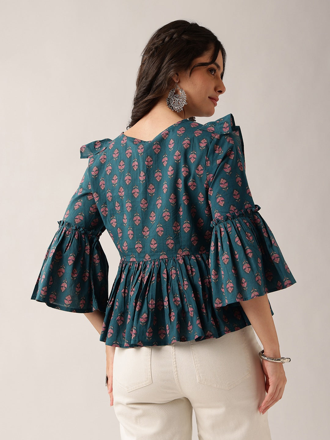 A woman wearing a teal green floral print blouse with ruffled sleeves and a peplum hem, paired with off-white trousers, shown from the back against a plain light gray studio background.
