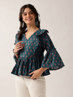 A woman modeling a dark teal green floral print blouse with ruffled shoulders and flared bell sleeves, paired with off-white pants, against a plain light beige background.