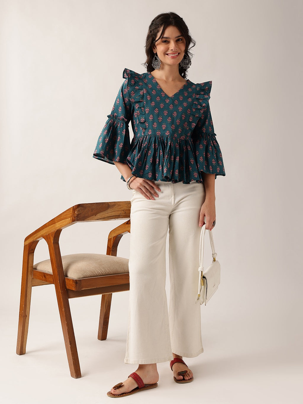 A woman stands in a studio wearing a dark teal floral ruffled blouse, off-white wide-leg cropped pants, and red sandals, holding a small white handbag next to a wooden chair.