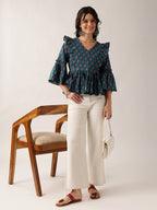 A woman stands in a studio wearing a dark teal floral ruffled blouse, off-white wide-leg cropped pants, and red sandals, holding a small white handbag next to a wooden chair.