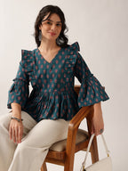 A woman wearing a dark teal ruffled blouse with a pink and green floral pattern, paired with cream-colored trousers, sitting on a wooden chair against a plain off-white background.