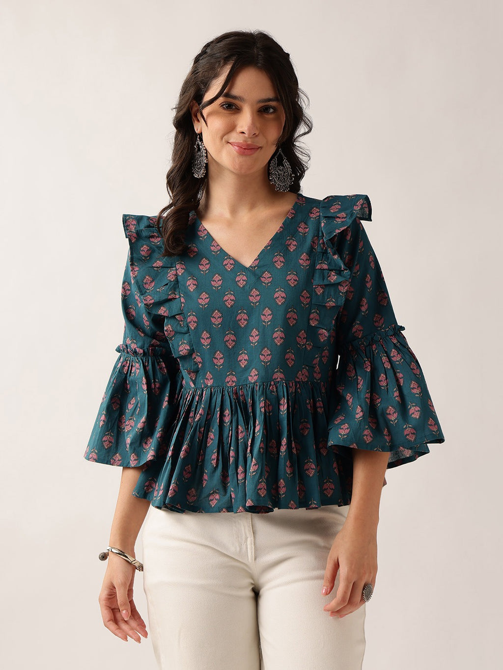 A woman wearing a dark teal blue blouse with a pink and green floral block print, ruffled shoulders, and flared bell sleeves, paired with off-white pants, against a plain light gray background.