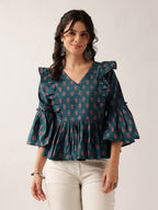 A woman wearing a dark teal blue blouse with a pink and green floral block print, ruffled shoulders, and flared bell sleeves, paired with off-white pants, against a plain light gray background.
