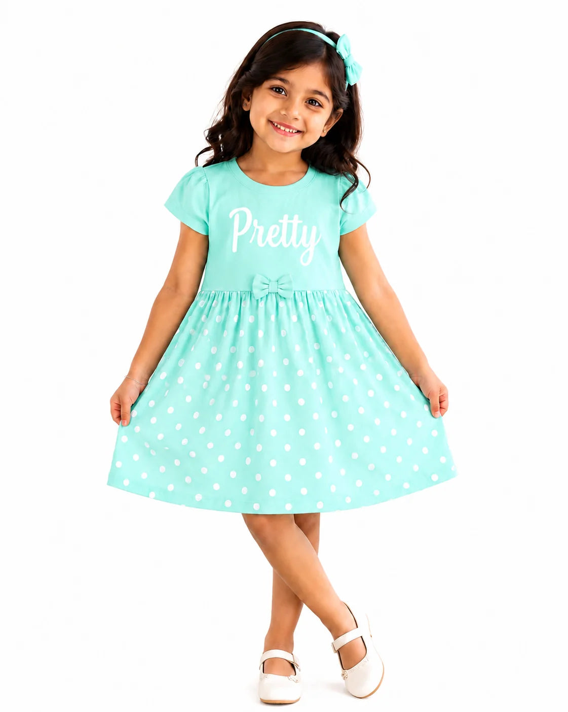 A young girl wearing a mint green short-sleeve dress with white polka dots and the word 'Pretty' printed in white on the chest, paired with a matching headband and white shoes, set against a plain white background.