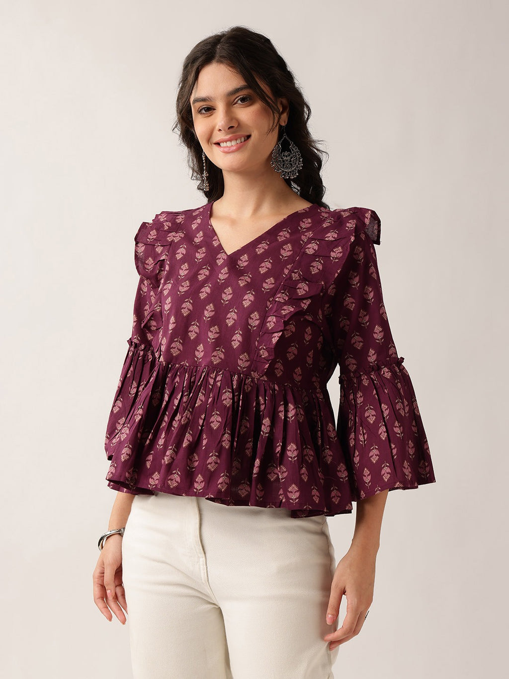 A woman wearing a dark purple floral print blouse with ruffled sleeves and a peplum hem, paired with off-white trousers, standing against a plain light gray background.