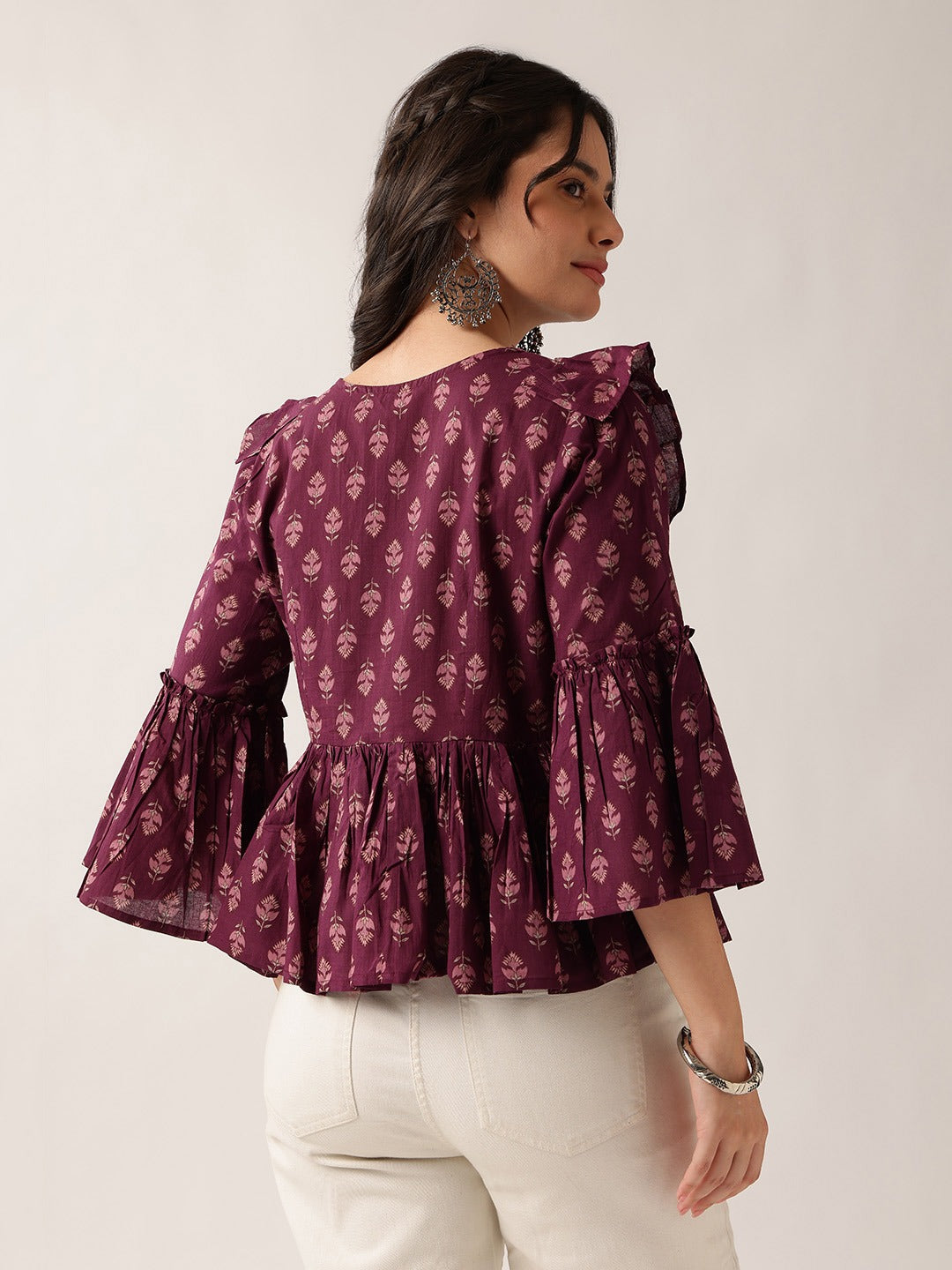 A woman wearing a deep purple long-sleeve blouse with a white floral block print and tiered ruffle details, paired with off-white trousers, shown from a three-quarter back view against a plain light background.