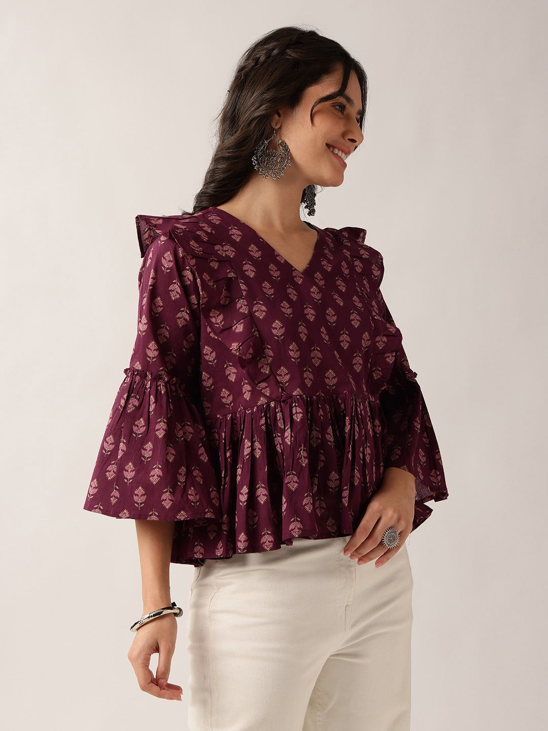 A woman wearing a deep purple V-neck blouse with a small white floral block print, ruffled shoulders, and flared bell sleeves, paired with white pants, set against a plain light background.