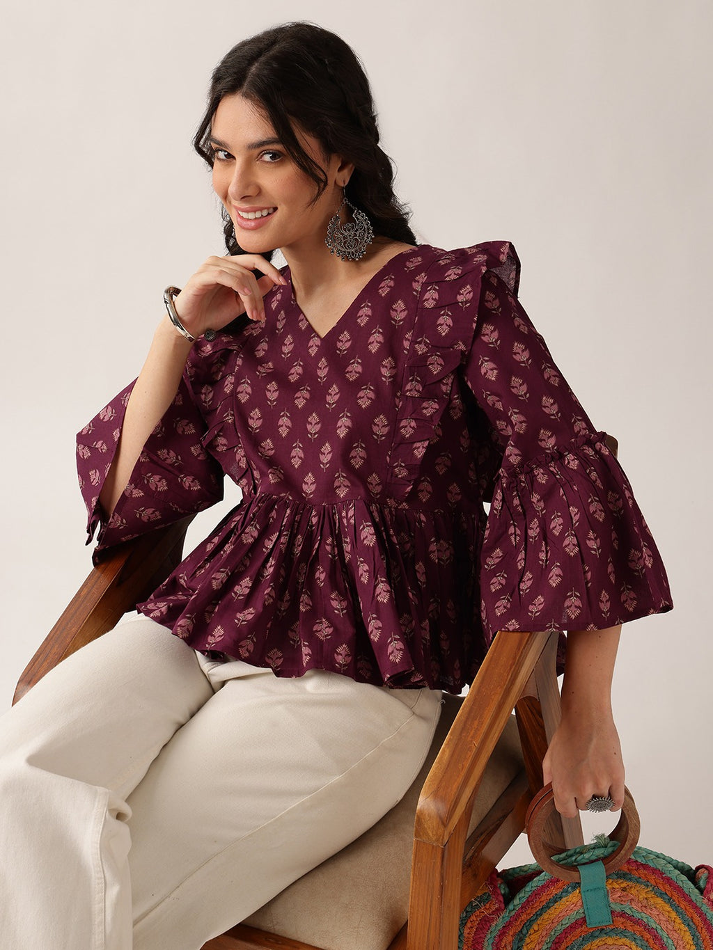 A woman seated in a wooden chair wearing a deep purple floral print blouse with flared bell sleeves and cream trousers, against a plain light background.