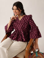 A woman seated in a wooden chair wearing a deep purple floral print blouse with flared bell sleeves and cream trousers, against a plain light background.