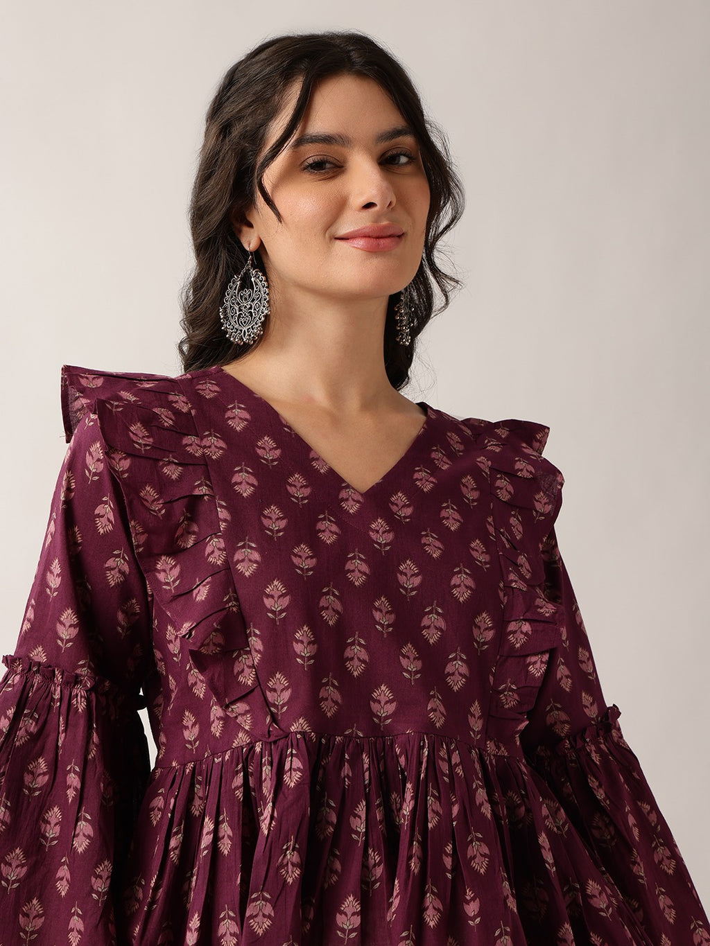 A woman wearing a deep purple long-sleeve dress with a repeating floral block print and ruffled details, paired with silver statement earrings, set against a plain light gray background.