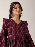 A woman wearing a deep purple long-sleeve dress with a repeating floral block print and ruffled details, paired with silver statement earrings, set against a plain light gray background.