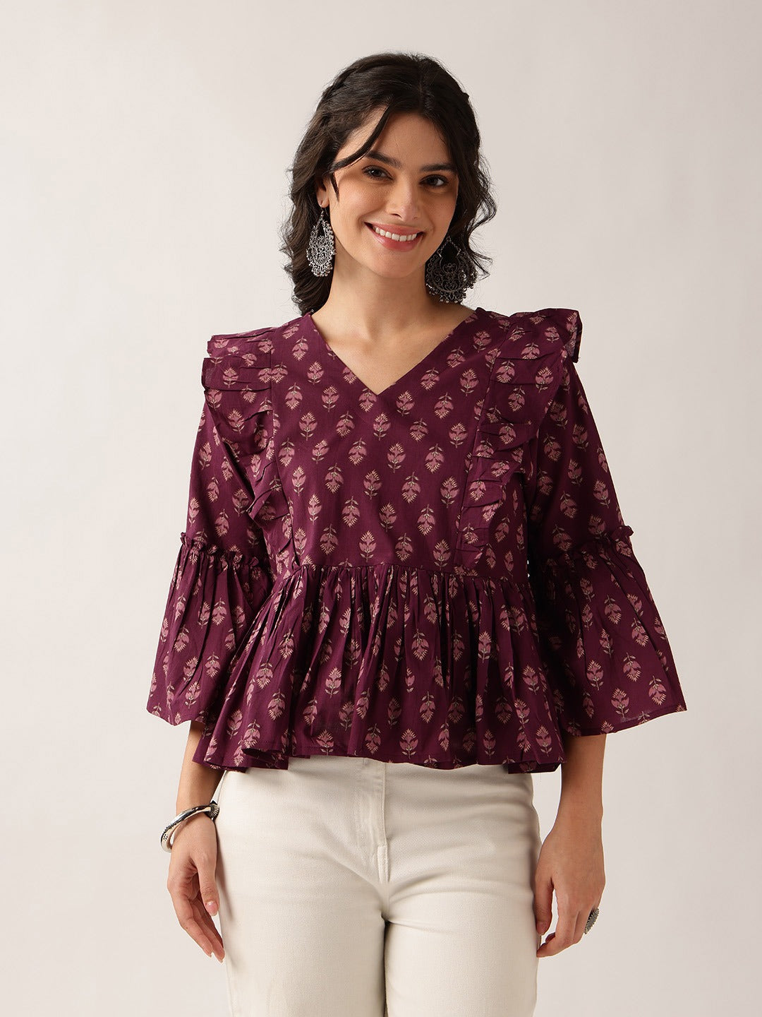 A woman wearing a dark purple, long-sleeved blouse with a small floral block print pattern, ruffled shoulders, and a gathered peplum waist, paired with off-white pants, against a plain light background.