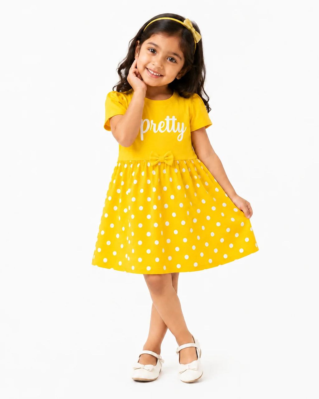 A young girl modeling a bright yellow short-sleeve dress with white polka dots and the word 'pretty' printed in white script across the chest. She is wearing a matching yellow headband and white Mary Jane shoes against a clean white background.