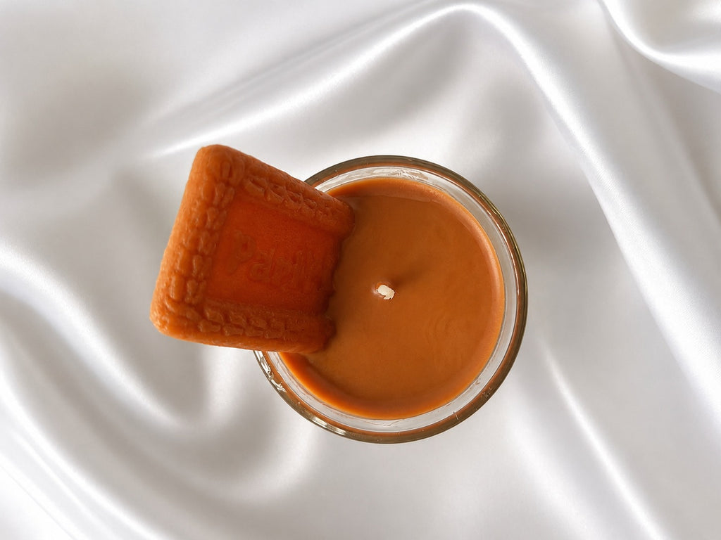 A top-down view of a candle in a clear glass jar filled with warm caramel-brown wax, with a textured orange rectangular block leaning against the rim, set on a draped white satin fabric background.