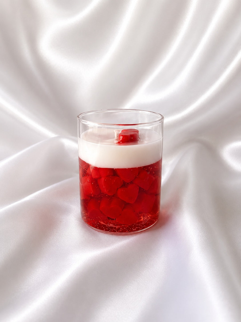A clear glass cup containing a layered red and white drink with red heart-shaped pieces suspended in the liquid, set against a draped white satin fabric background.
