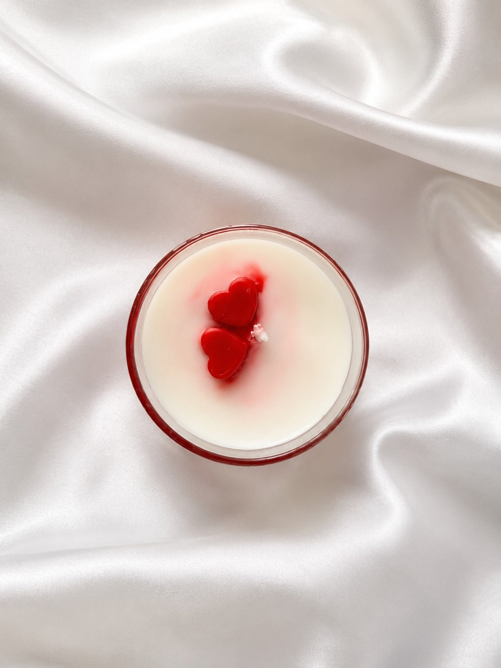A round glass candle with creamy off-white wax and two red heart-shaped wicks, set on a draped white satin fabric background.