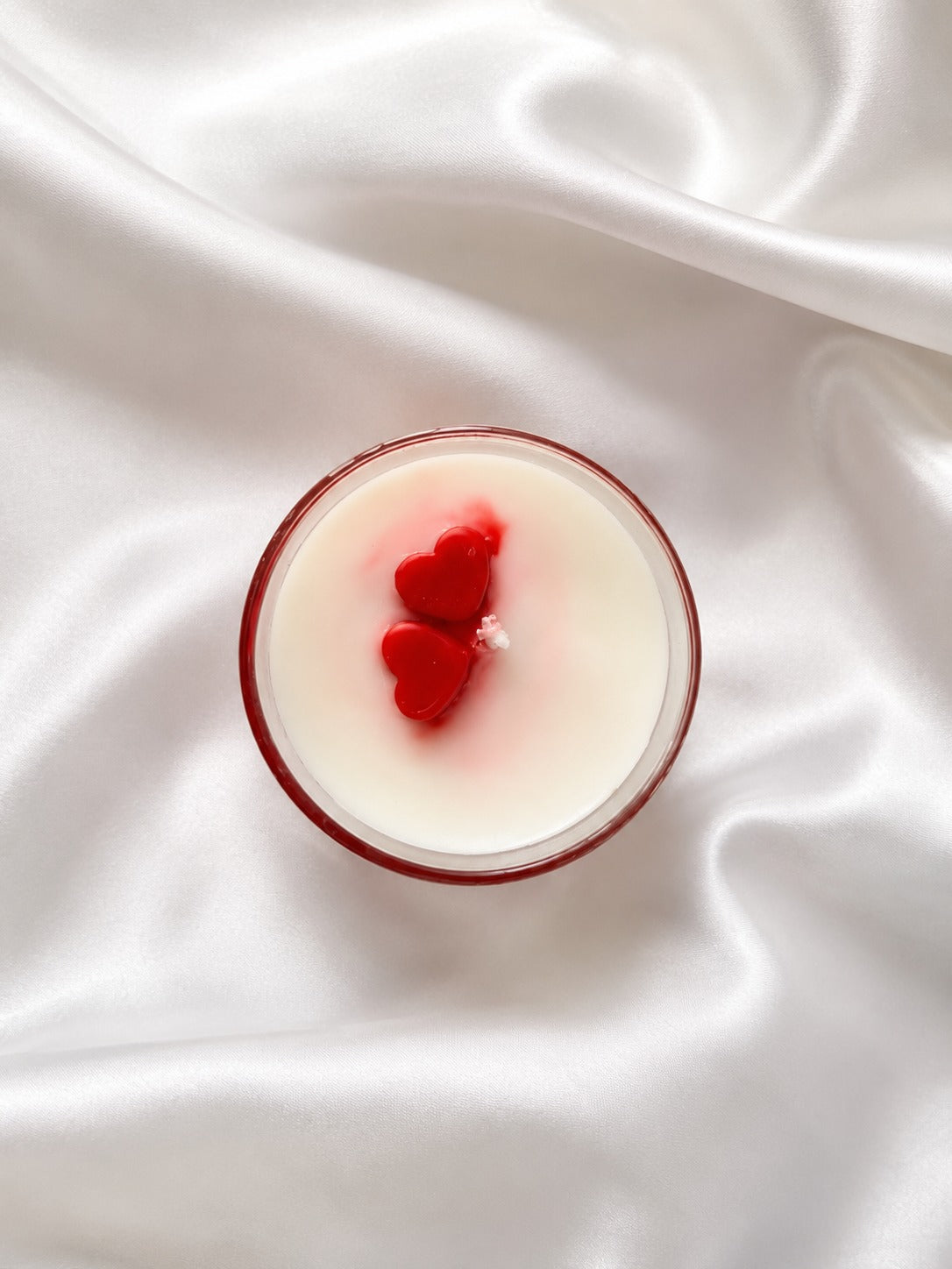 A round glass candle with creamy off-white wax and two red heart-shaped wicks, set on a draped white satin fabric background.