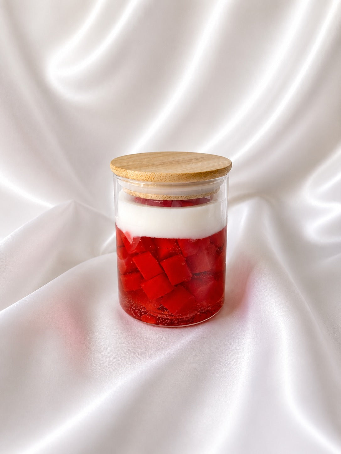 A clear glass jar with a light wood lid, containing red fruit cubes and a thick white layer, set against a draped white satin fabric background.