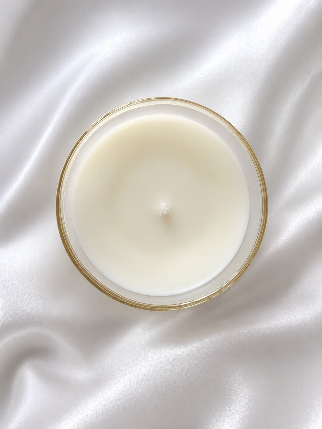 A top-down view of a single-wick candle in a clear glass jar with a thin gold rim, set against a draped white satin fabric background.