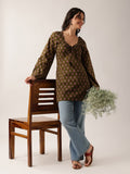 A woman models a brown long-sleeve tunic top with a small beige floral pattern, paired with light blue jeans and brown sandals, standing next to a wooden chair and holding a bouquet of baby's breath.