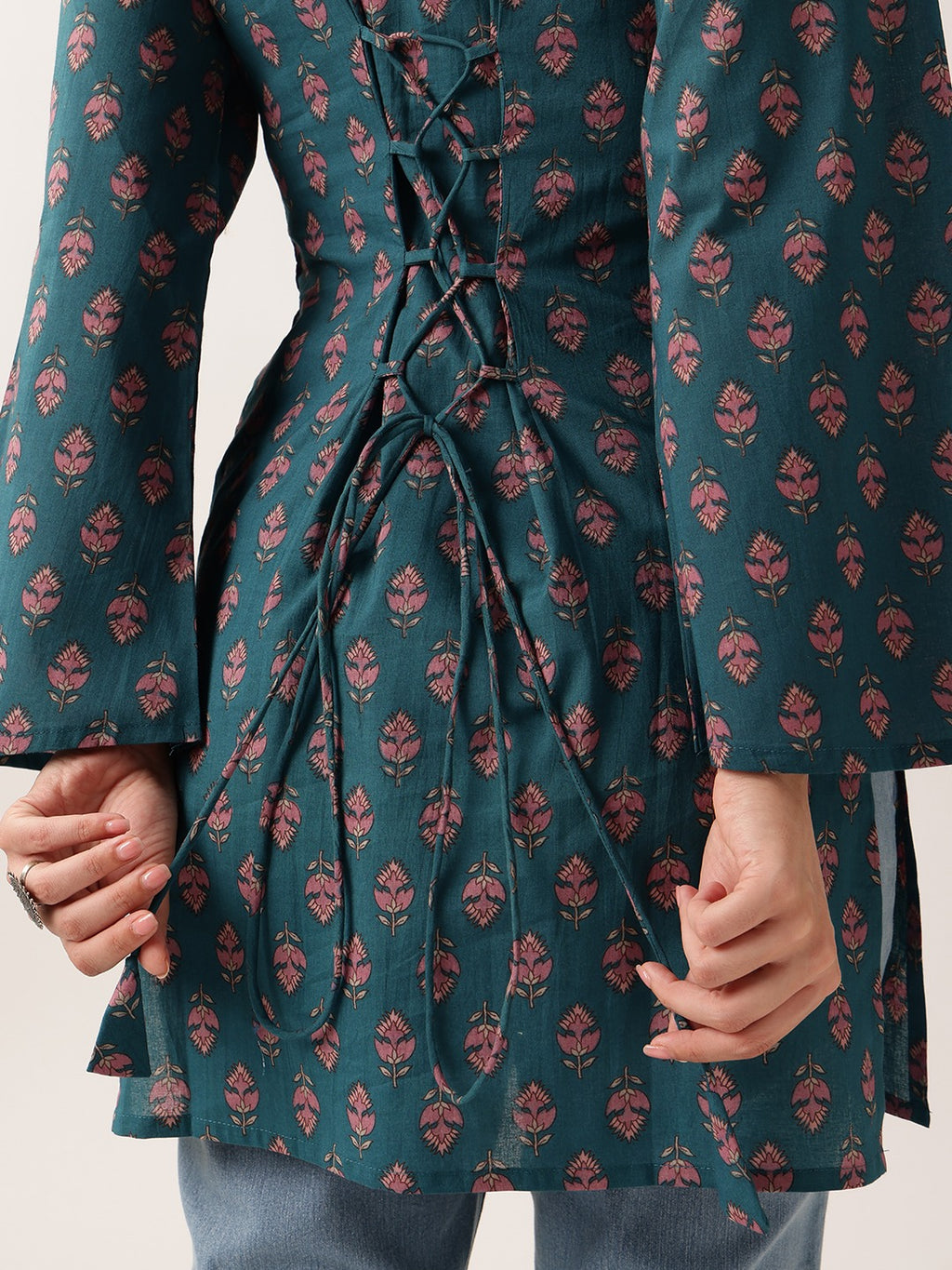 A close-up of a teal green tunic with a pink floral block print and a lace-up front, worn by a model with blue jeans visible at the bottom.