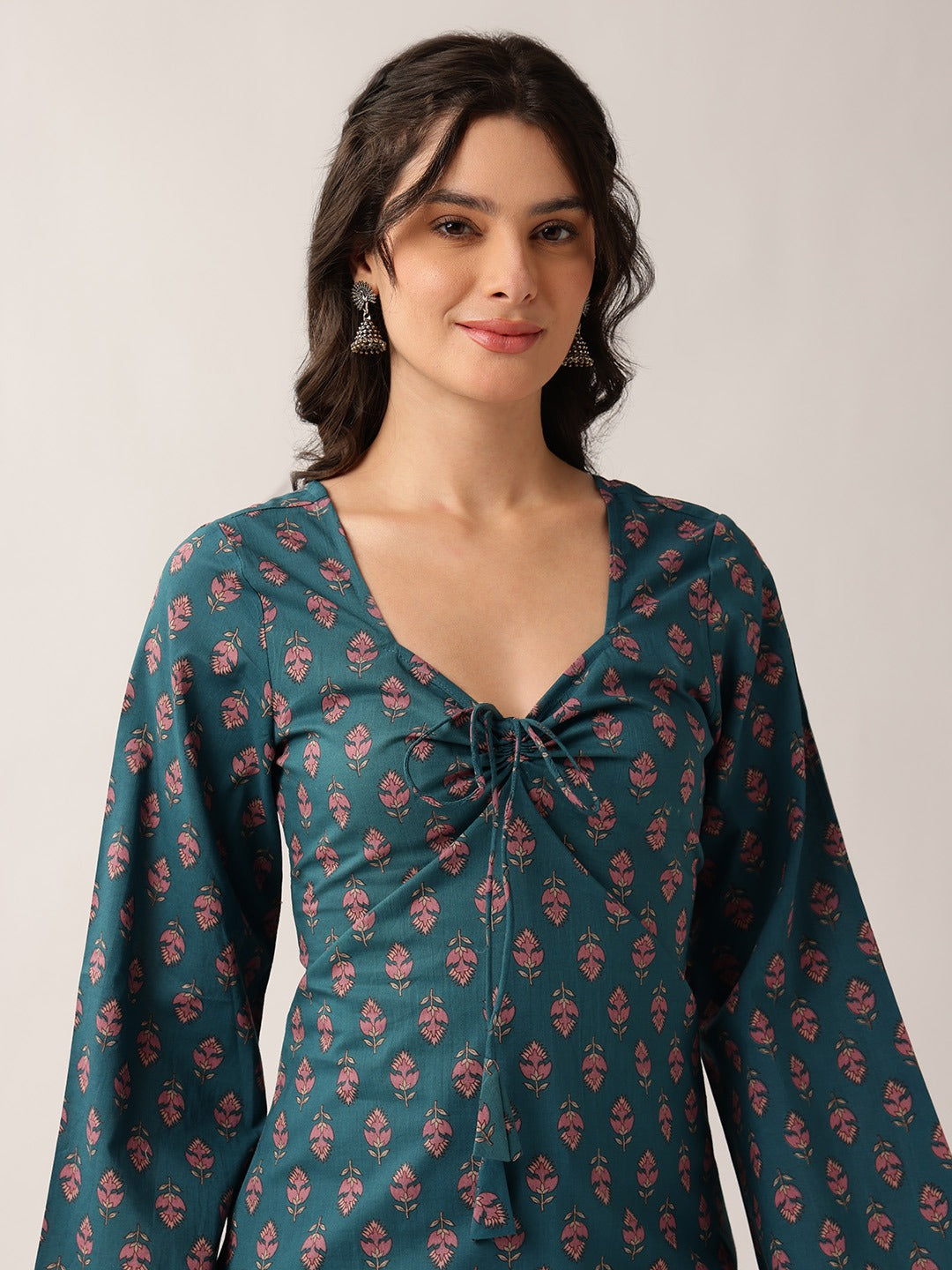 A woman wearing a teal long-sleeve kurta with a pink floral block print and a front tie detail, photographed against a neutral light gray background.