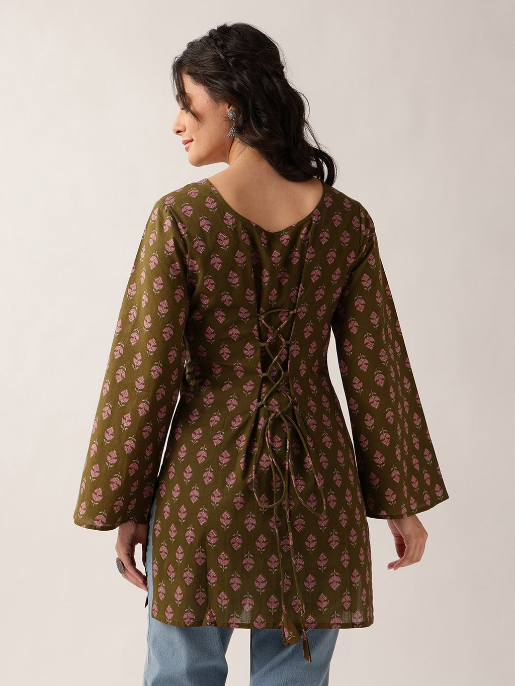 A woman shown from the back wearing a brown tunic-style top with a pink floral block print and lace-up back details, paired with light blue jeans against a plain light background.