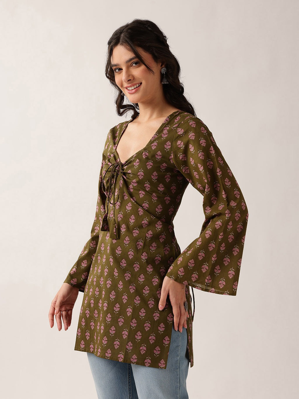 A woman modeling a brown long-sleeve tunic top with a pink floral block print pattern, featuring a tie-front detail and flared bell sleeves, paired with light blue jeans against a plain light gray background.