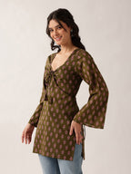 A woman modeling a brown long-sleeve tunic top with a pink floral block print pattern, featuring a tie-front detail and flared bell sleeves, paired with light blue jeans against a plain light gray background.