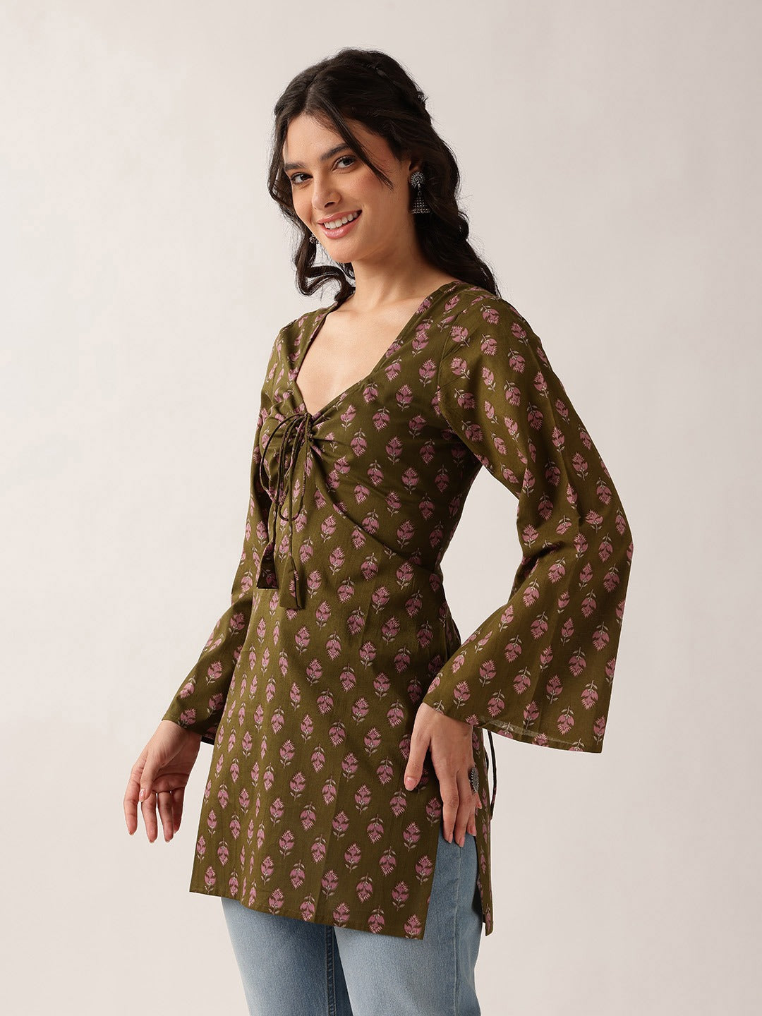 A woman modeling a brown long-sleeve tunic top with a pink floral block print pattern, featuring a tie-front detail and flared bell sleeves, paired with light blue jeans against a plain light gray background.