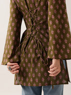 A close-up of a person wearing an olive green tunic top with a pink floral block print and lace-up back detail, paired with blue jeans.