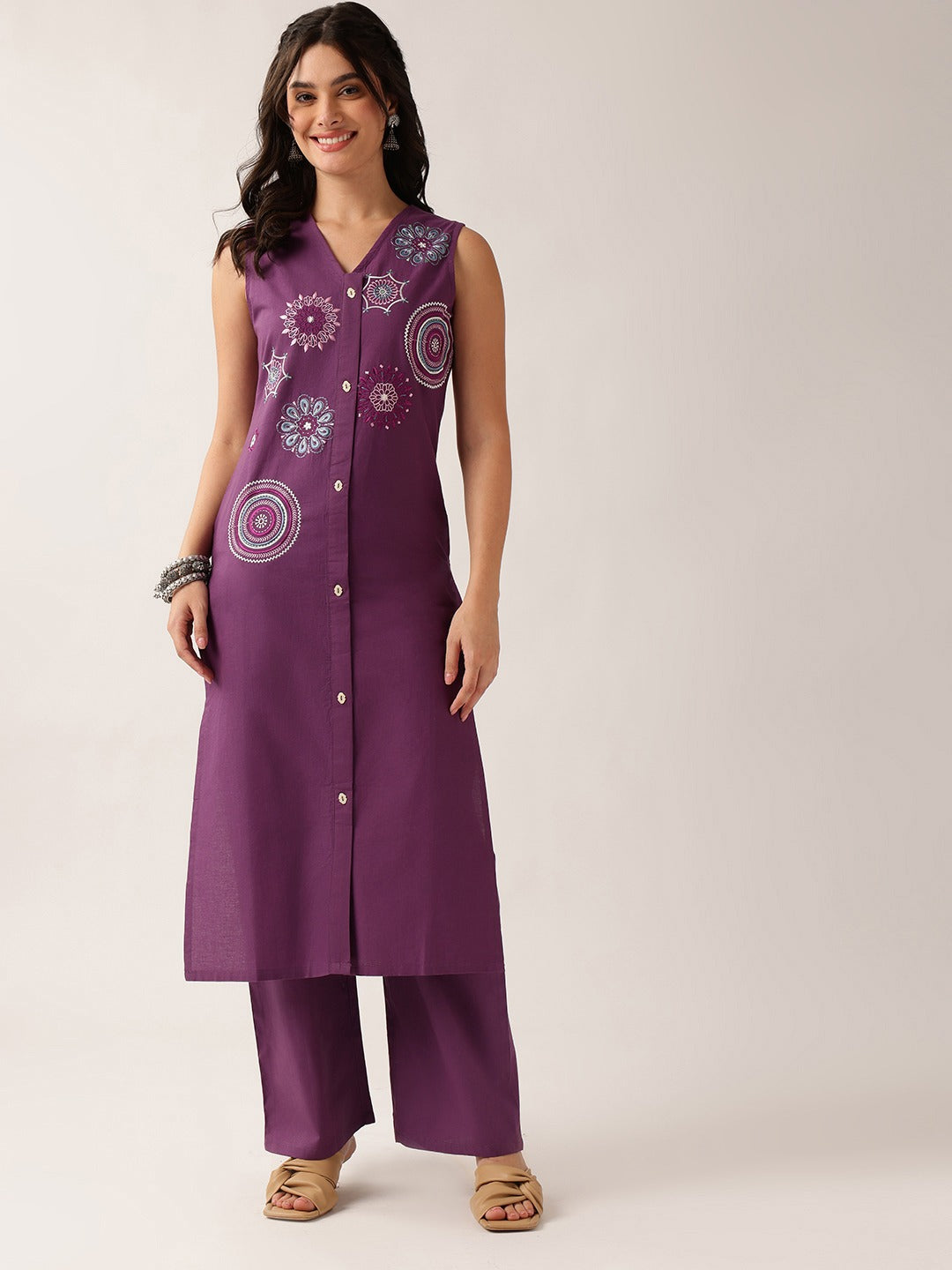 A woman modeling a sleeveless purple button-front tunic with matching wide-leg pants. The tunic features white and light purple floral and circular embroidered patterns on the front.