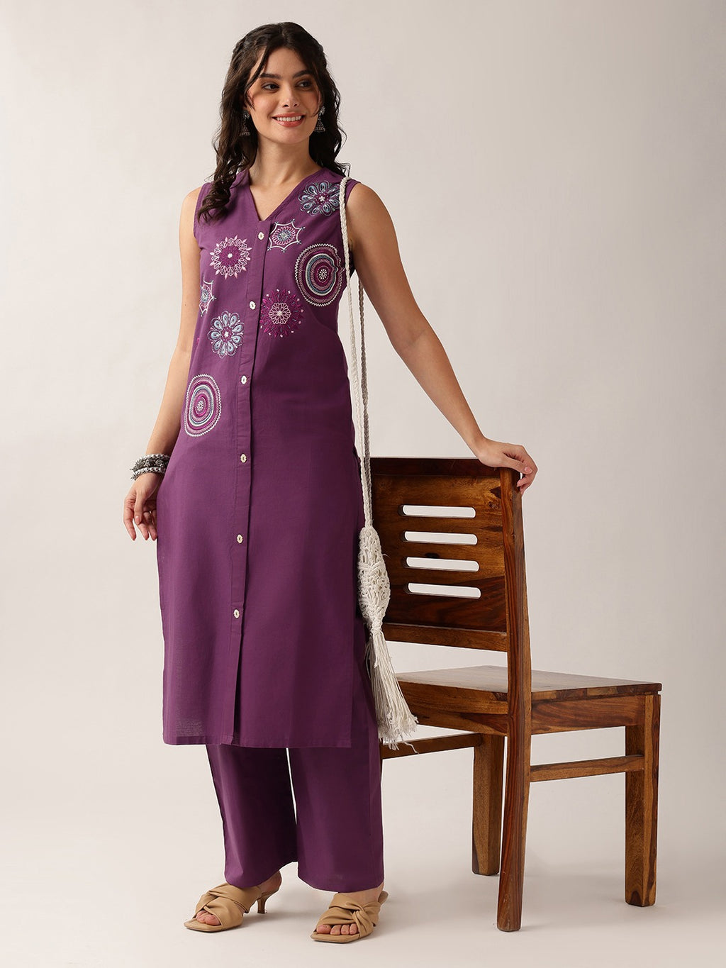 A woman models a purple sleeveless kurta and matching wide-leg pants set with white floral embroidery, standing next to a wooden chair against a light beige background.