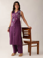 A woman models a purple sleeveless kurta and matching wide-leg pants set with white floral embroidery, standing next to a wooden chair against a light beige background.