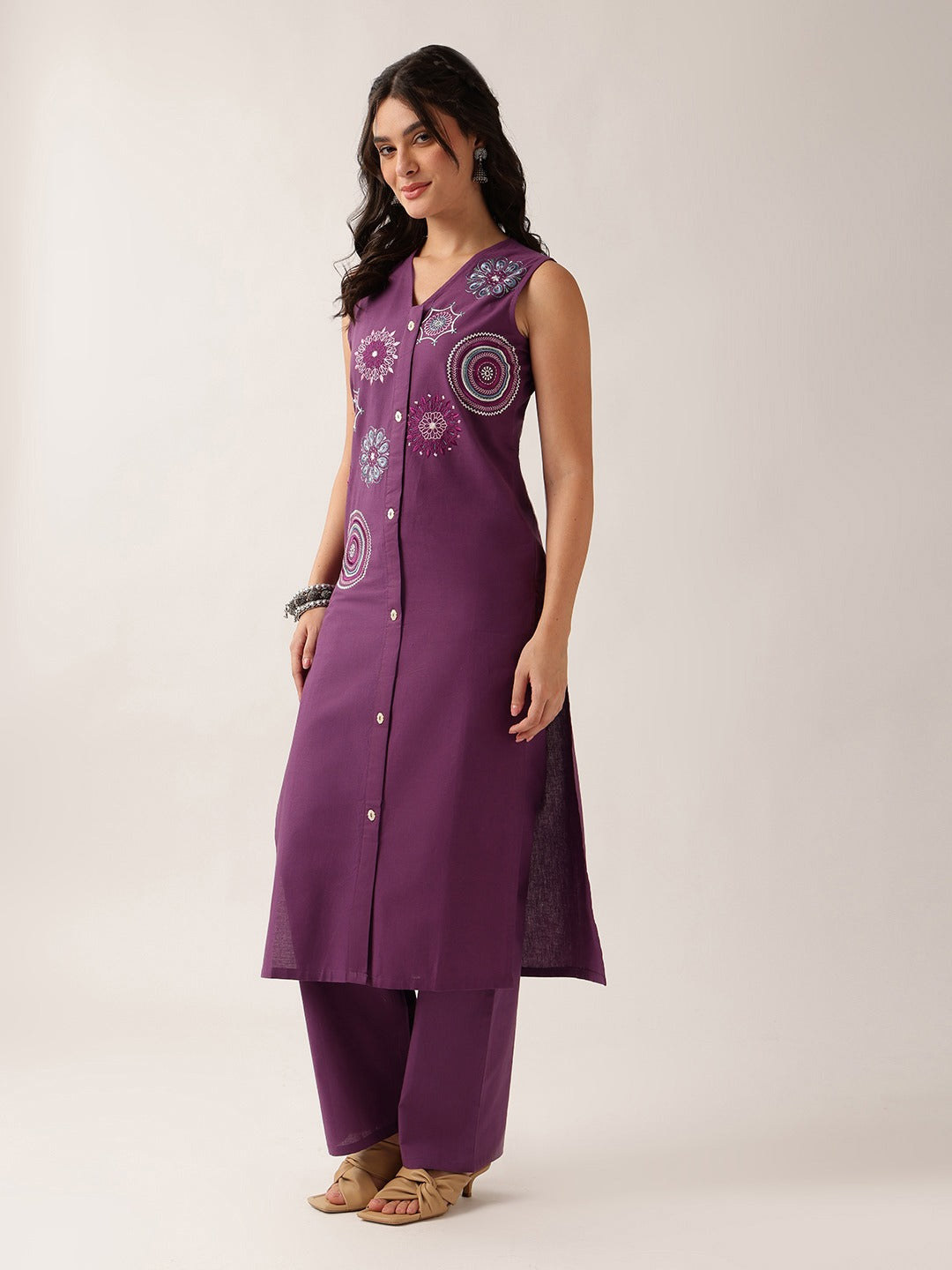 A woman modeling a sleeveless purple kurta with circular embroidered patterns on the chest and matching wide-leg pants, photographed against a plain light gray studio background.