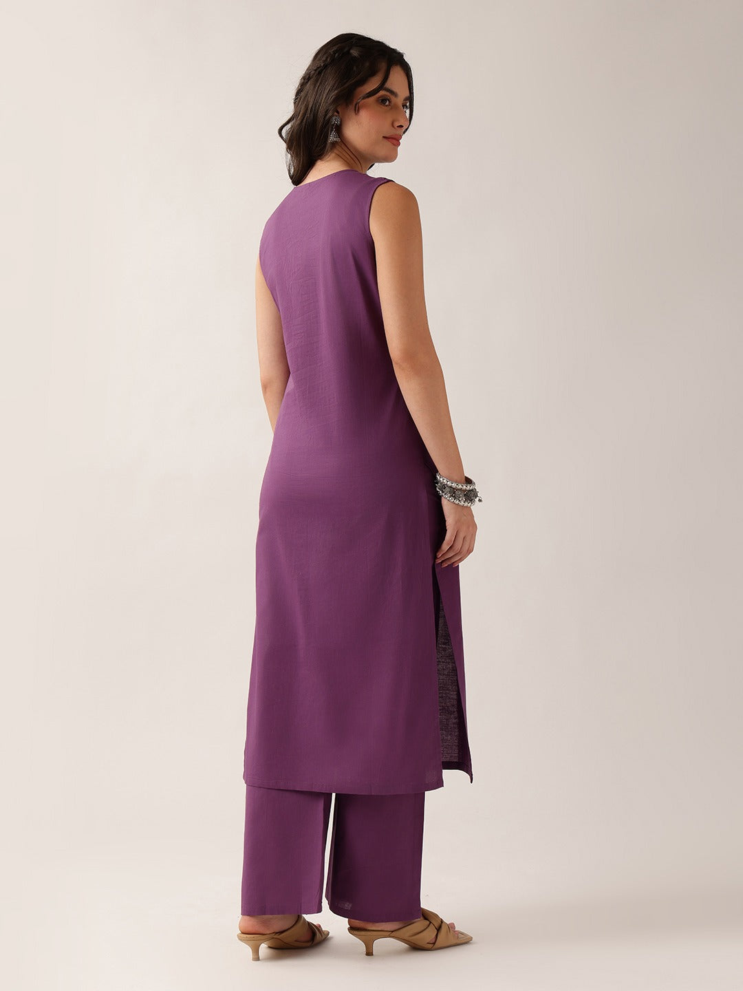 A woman modeling a sleeveless purple kurta with matching wide-leg trousers, shown from a rear three-quarter angle against a plain light beige studio background.