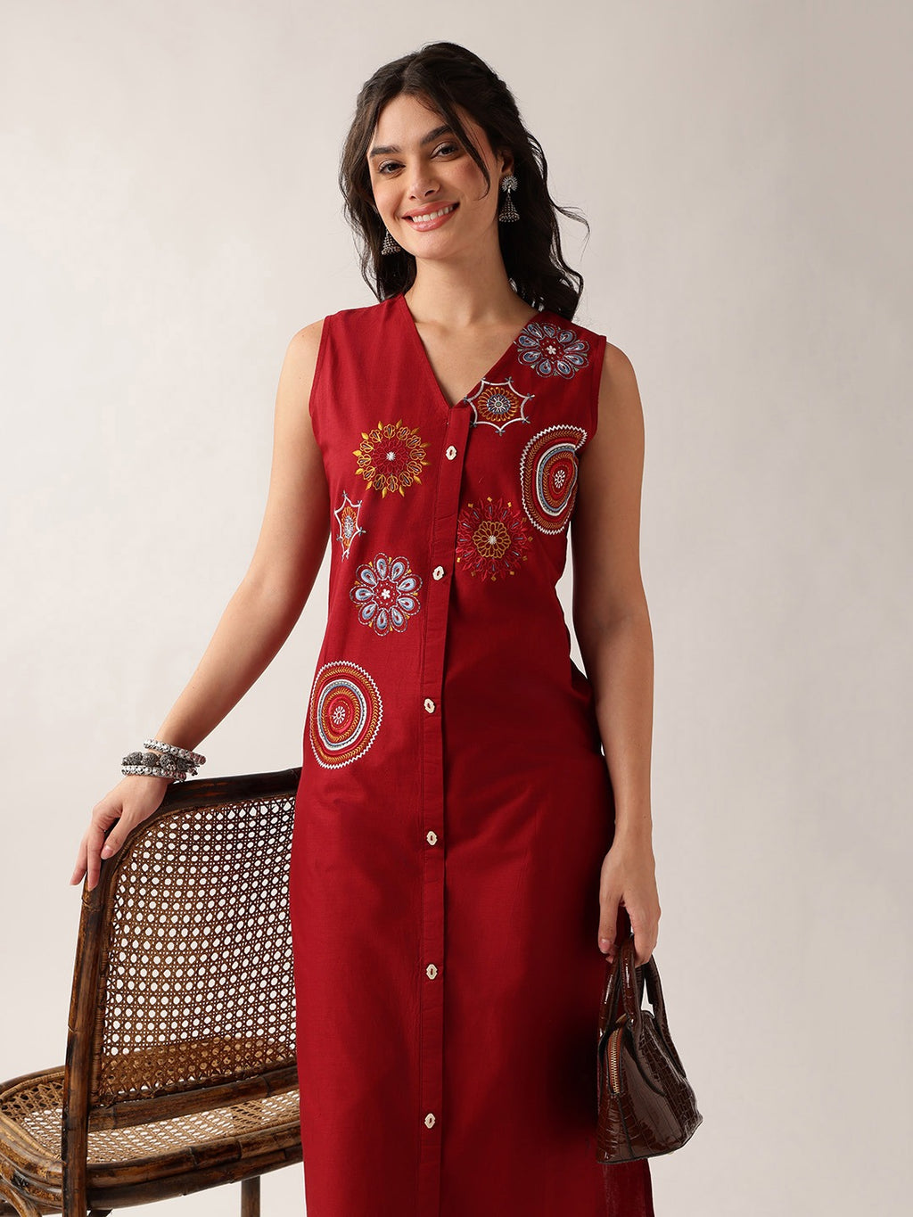 A woman models a sleeveless red button-front dress with colorful circular and floral embroidered motifs on the front. She is standing next to a woven rattan chair against a plain light beige studio background, holding a small brown handbag.