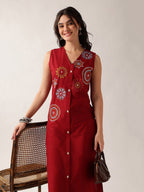 A woman models a sleeveless red button-front dress with colorful circular and floral embroidered motifs on the front. She is standing next to a woven rattan chair against a plain light beige studio background, holding a small brown handbag.