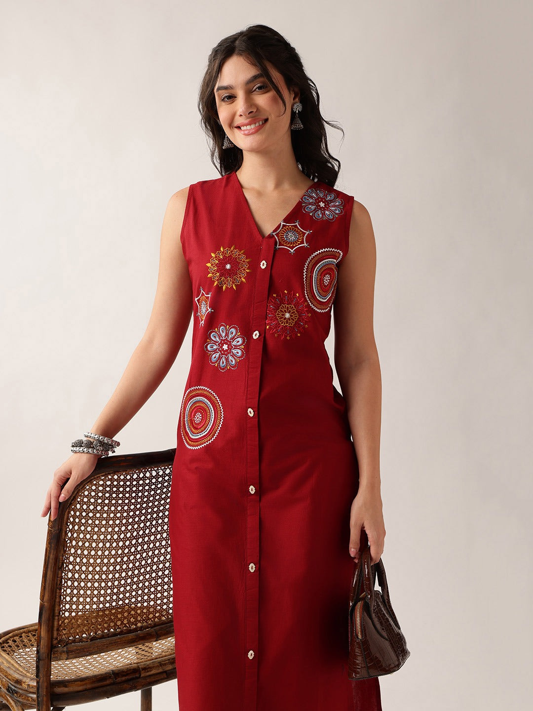 A woman models a sleeveless red button-front dress with colorful circular and floral embroidered motifs on the front. She is standing next to a woven rattan chair against a plain light beige studio background, holding a small brown handbag.