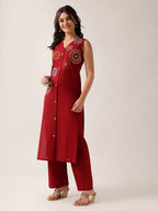 A woman modeling a sleeveless red kurta with colorful circular embroidery on the chest and matching red wide-leg pants, photographed against a plain light beige studio background.