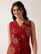 A woman wearing a sleeveless red button-up top with colorful embroidered circular and floral motifs, photographed against a plain light background.
