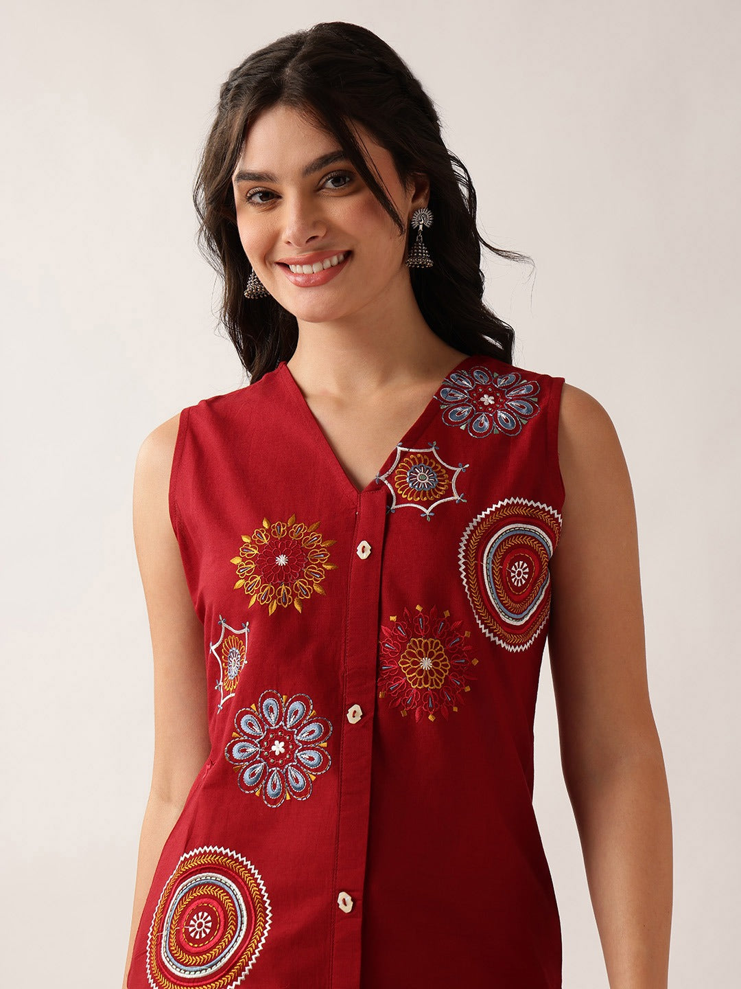 A woman wearing a sleeveless red button-up top with colorful embroidered circular and floral motifs, photographed against a plain light background.